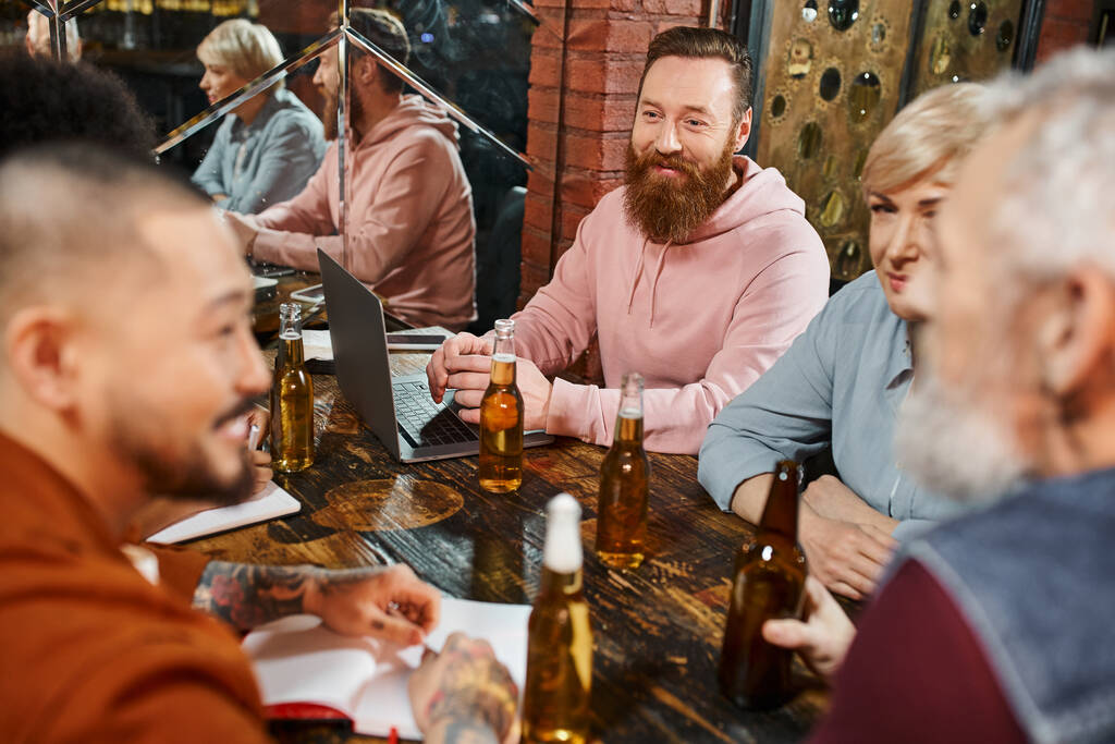 χαμογελαστοί πολυεθνικοί συνάδελφοι που μιλούν κοντά σε μπουκάλια μπύρας και laptop κατά τη διάρκεια συνάντησης σε παμπ μετά τη δουλειά - Φωτογραφία, εικόνα