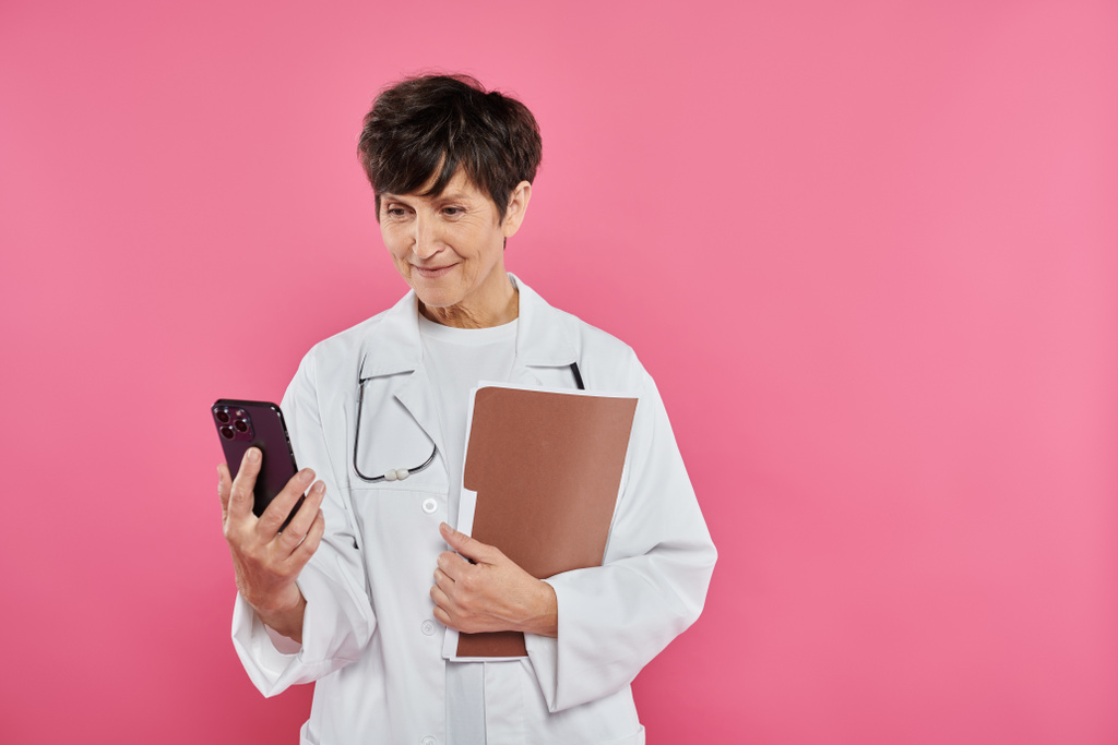 volwassen oncoloog, vrouwelijke arts holding folder, met behulp van smartphone, borstkanker bewustzijn concept - Foto, afbeelding