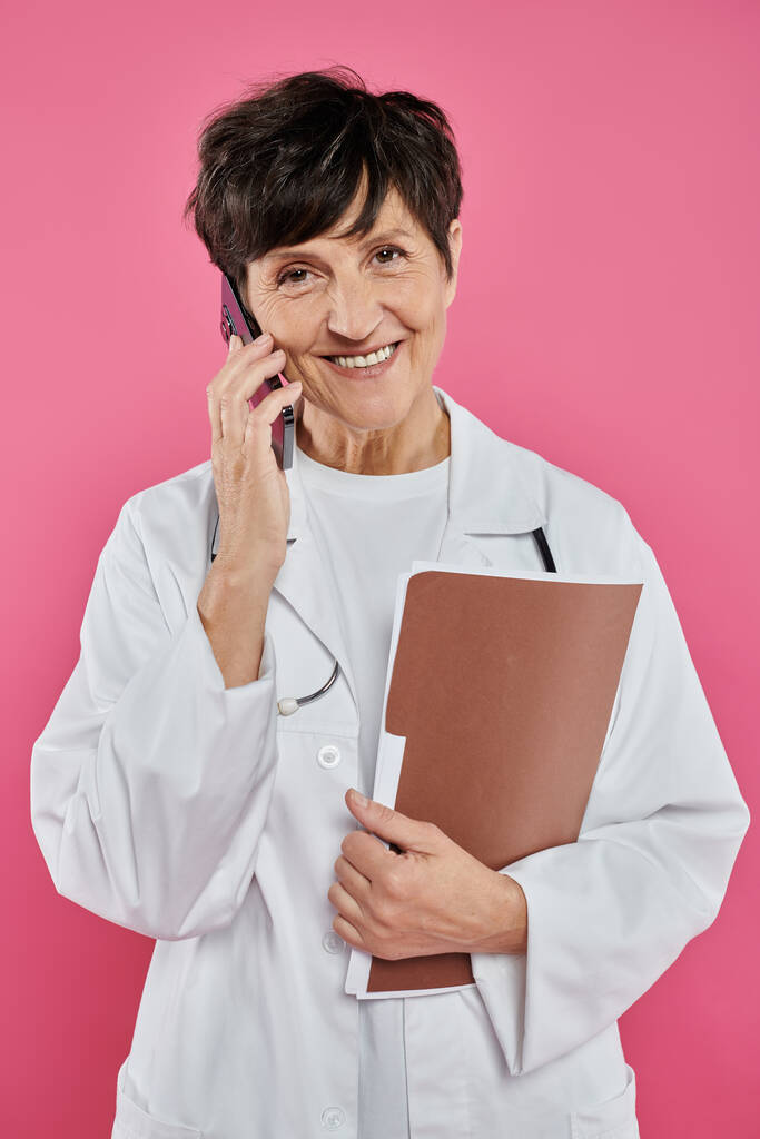 mature oncologist, female doctor holding folder and talking on smartphone, breast cancer awareness - Photo, Image