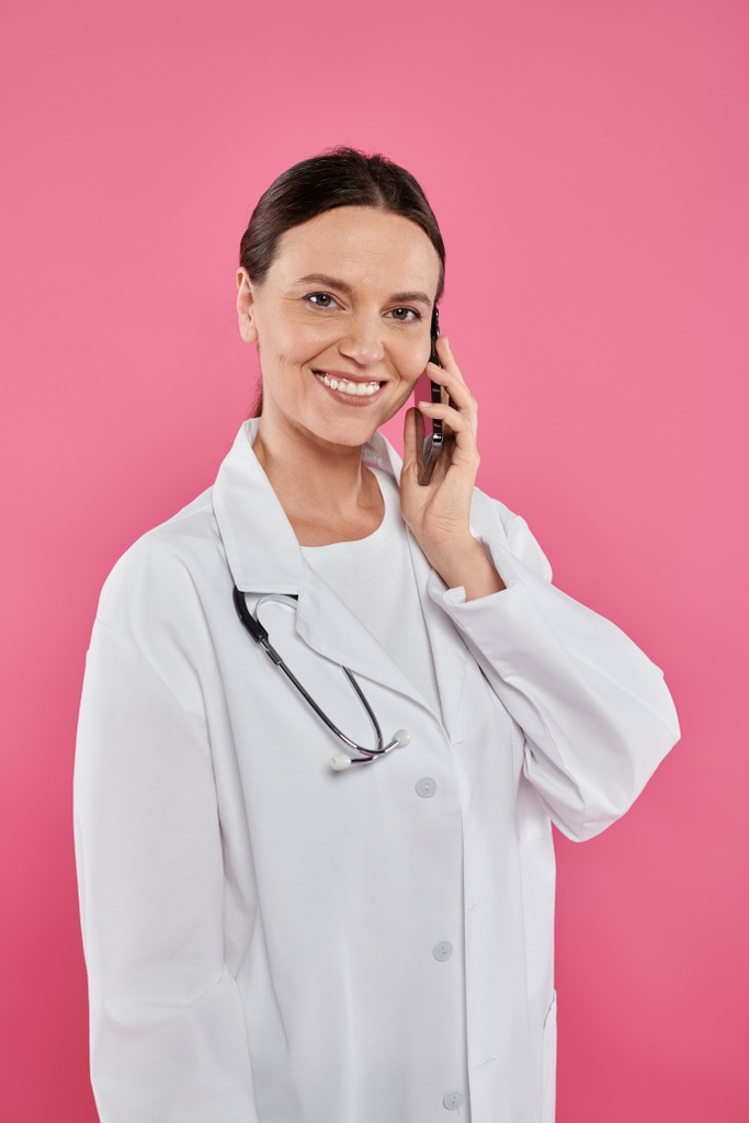 ピンクで隔離されたスマートフォンで話す笑顔の女性医師,乳がんの概念 - 写真・画像