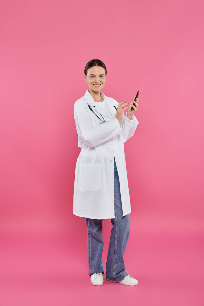 smiling doctor in white coat holding smartphone and looking at camera on pink, breast cancer concept - Photo, Image