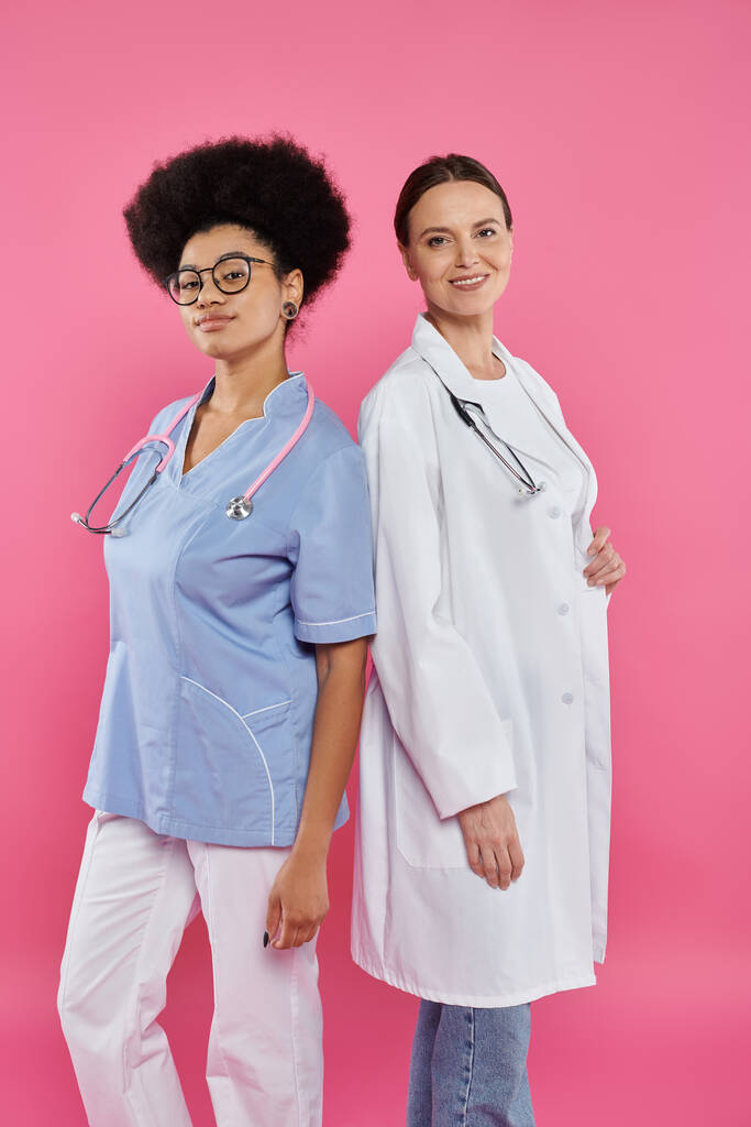 smiling multiethnic doctors looking at camera isolated on pink, breast cancer concept - Photo, Image