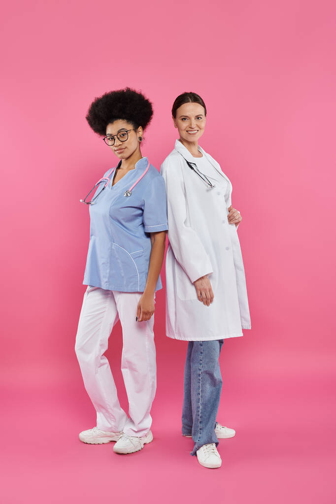 alegres médicos interracial mirando a la cámara en rosa, concepto de conciencia del cáncer de mama - Foto, Imagen