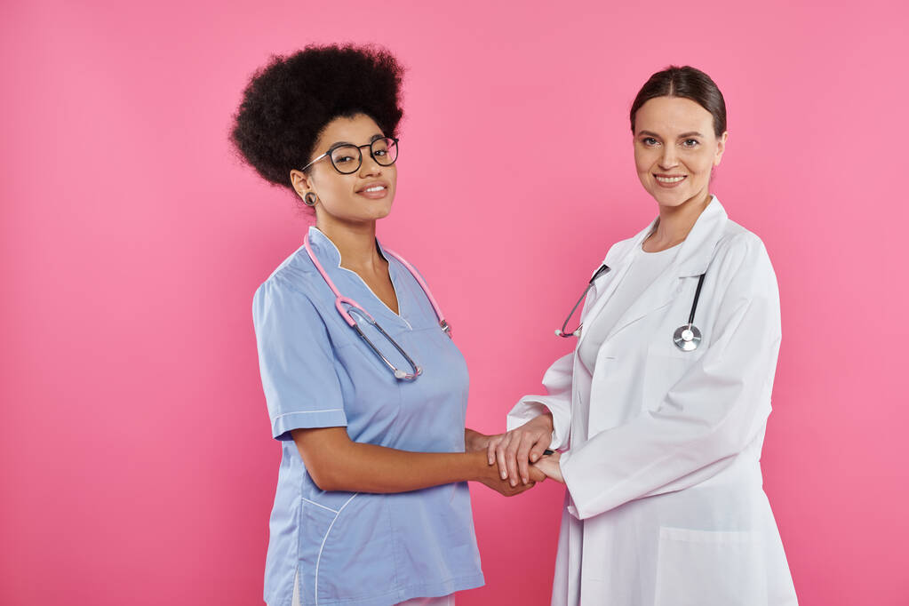 Lächeln multiethnische Ärzte Händchen haltend und isoliert auf rosa, Brustkrebs Bewusstsein - Foto, Bild