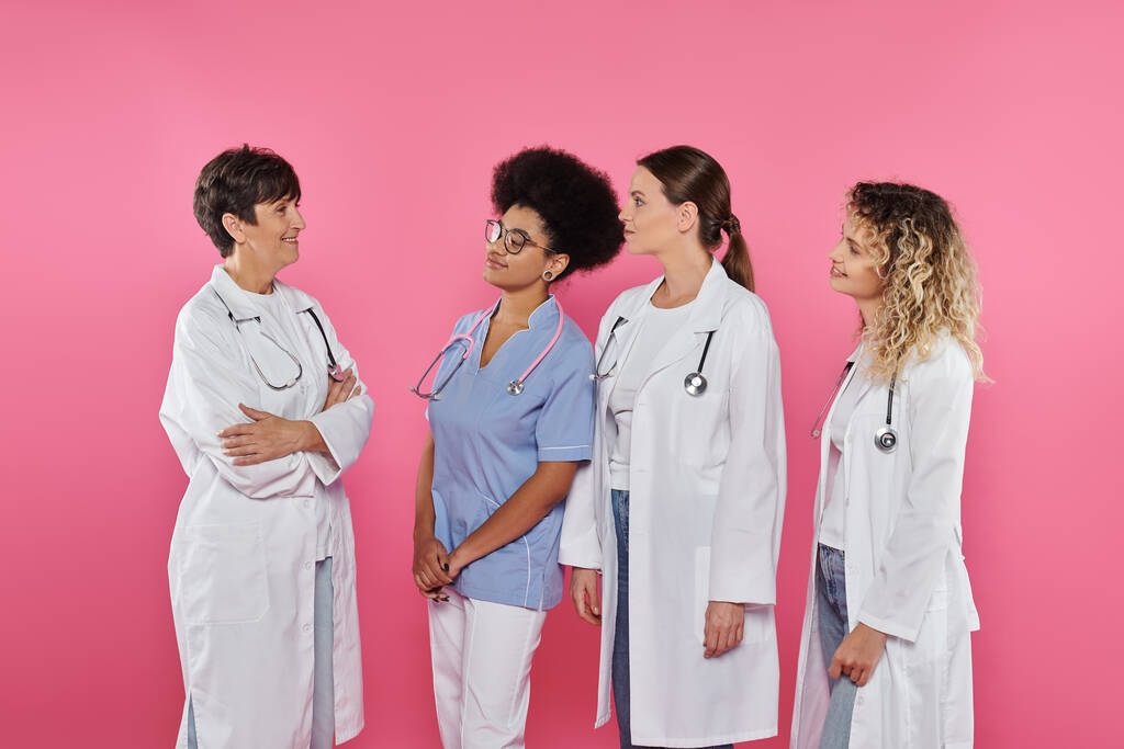 multiethnische Ärzte schauen lächelnde Kollegen an, die die Arme isoliert auf rosa, Brustkrebs Monat kreuzen - Foto, Bild