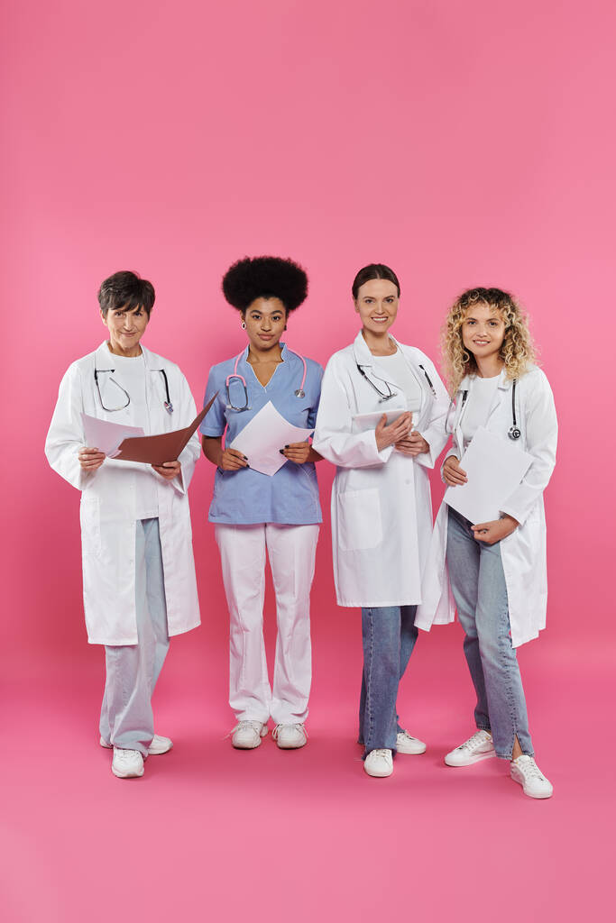 uśmiechnięci międzyrasowi lekarze trzymający papierowe teczki i cyfrową tabletkę na różowym, miesiącu raka piersi - Zdjęcie, obraz