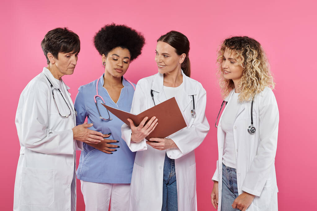 glimlachende arts met papieren map in de buurt van multi-etnische collega 's geïsoleerd op roze, borstkanker - Foto, afbeelding
