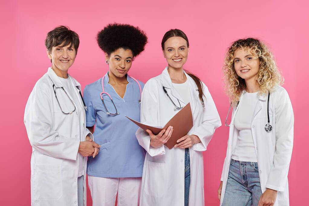 счастливые многоэтнические врачи с бумажной папкой и глядя на камеру изолированы на розовый, рак молочной железы - Фото, изображение