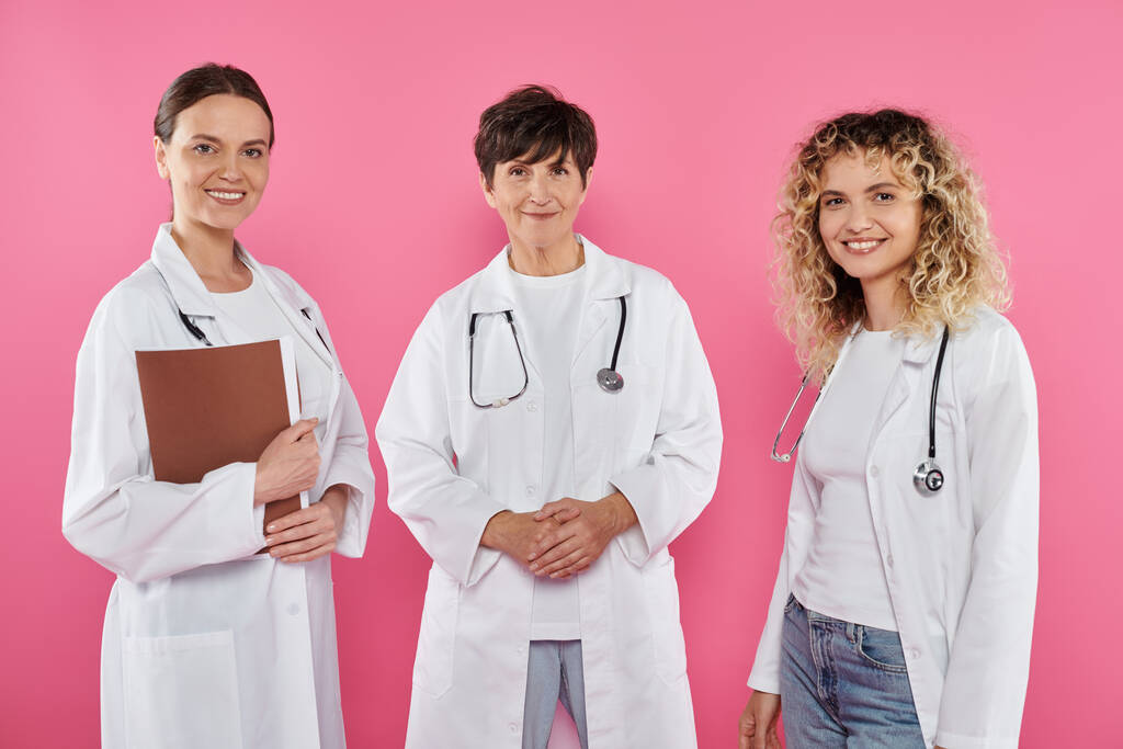 Lächelnde Ärztinnen in weißen Mänteln, die in die Kamera schauen, isoliert auf rosa, Brustkrebs-Bewusstsein - Foto, Bild