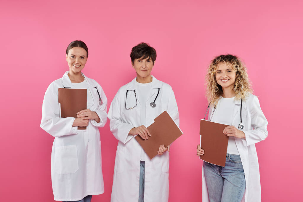 веселые врачи держат бумажные папки, стоя изолированно на розовой концепции рака груди - Фото, изображение
