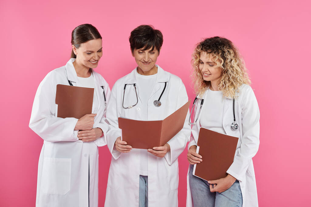 smiling doctors looking at paper folder together isolated on pink, breast cancer awareness month - Photo, Image