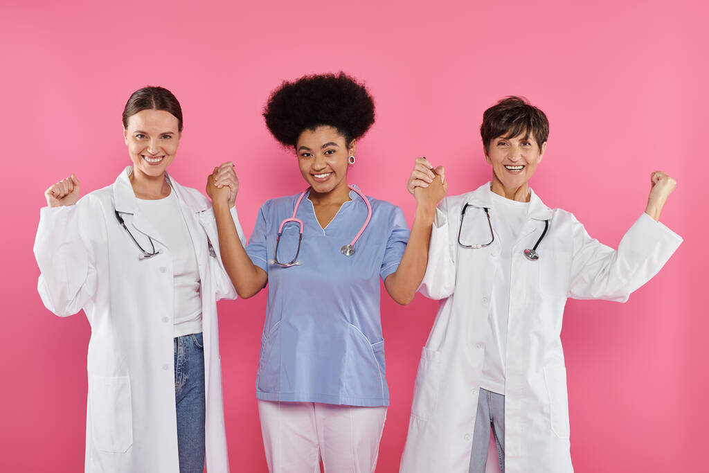 médicos oncólogos multiétnicos emocionados tomados de las manos aislados en rosa, mes del cáncer de mama - Foto, imagen