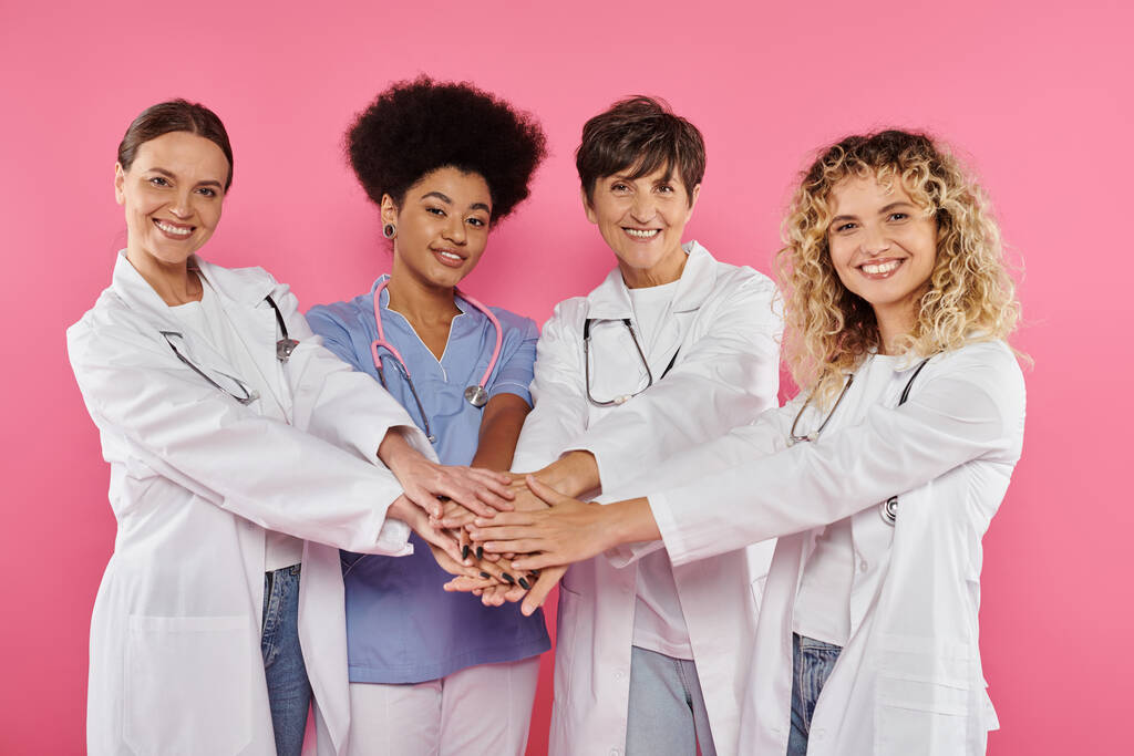 ομάδα χαρούμενων πολυεθνικών ογκολόγων κρατώντας τα χέρια απομονώνονται σε ροζ, έννοια του καρκίνου του μαστού - Φωτογραφία, εικόνα