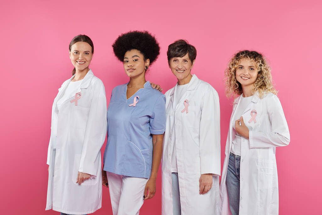 sorridenti medici interrazziali con nastri che guardano la fotocamera isolata sul rosa, concetto di cancro al seno - Foto, immagini