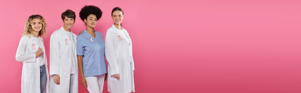 smiling multiethnic doctors with ribbons on coats standing isolated on pink, banner, breast cancer - Photo, Image