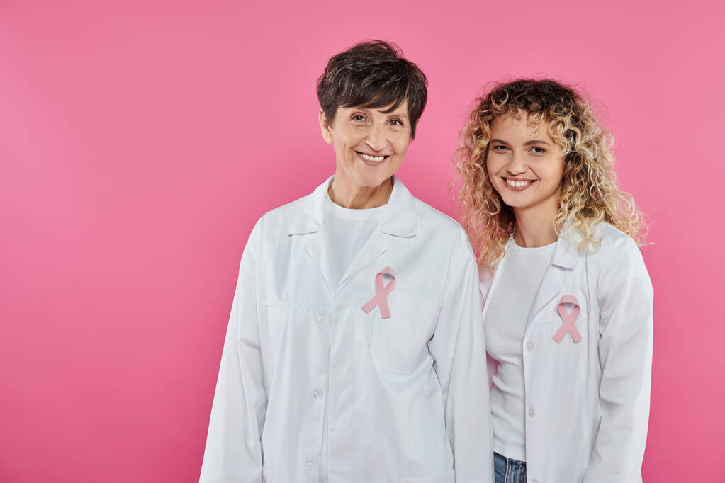 cheerful oncologists with ribbons on white coats standing isolated on pink, breast cancer concept - Photo, Image