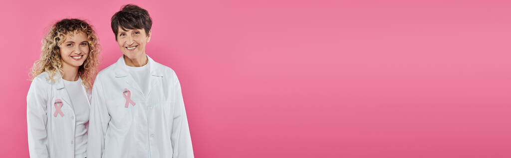 χαμογελαστοί γιατροί με κορδέλες σε λευκά παλτά που απομονώνονται σε ροζ, banner, έννοια του καρκίνου του μαστού - Φωτογραφία, εικόνα