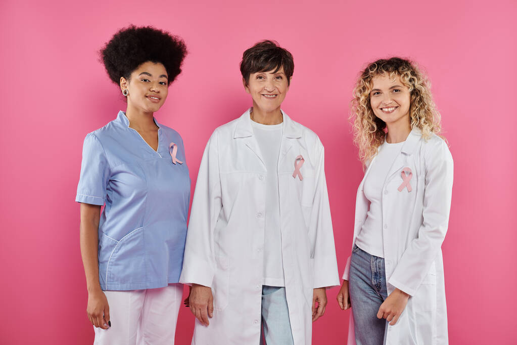 positive interracial doctors with ribbons of breast cancer standing together on pink background - Photo, Image