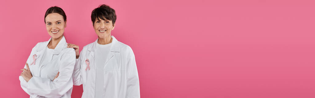 усміхнені лікарі зі стрічками на білих пальто позують ізольовані на рожевому, концепції раку молочної залози, банер - Фото, зображення