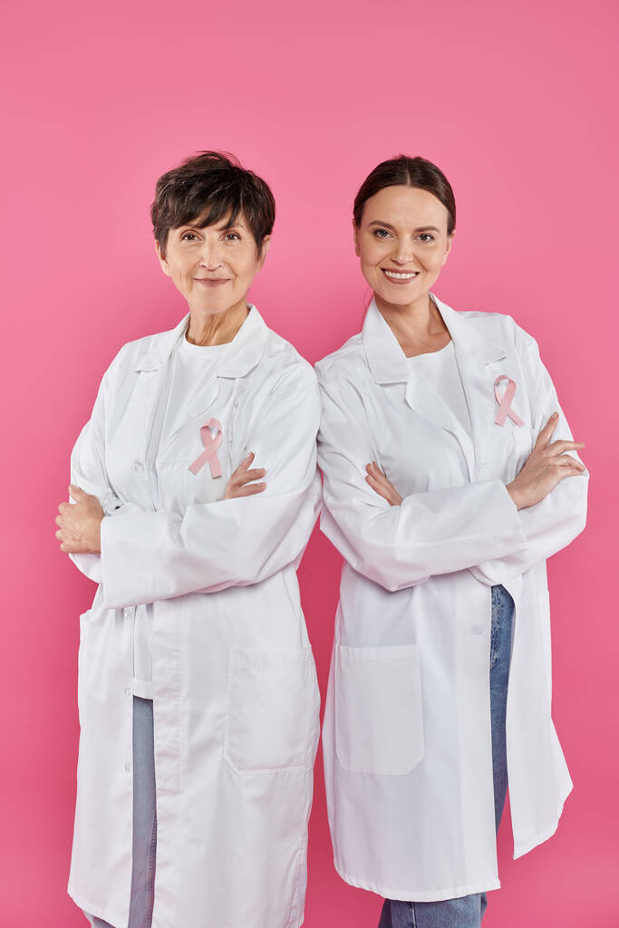 smiling female doctors with ribbons crossing arms isolated on pink, breast cancer awareness - Photo, Image
