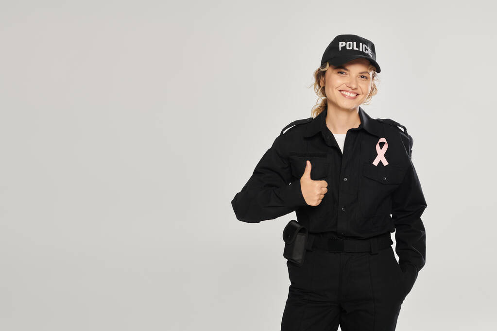 smiling policewoman with pink ribbon showing thumb up isolated on grey, breast cancer month - Photo, Image