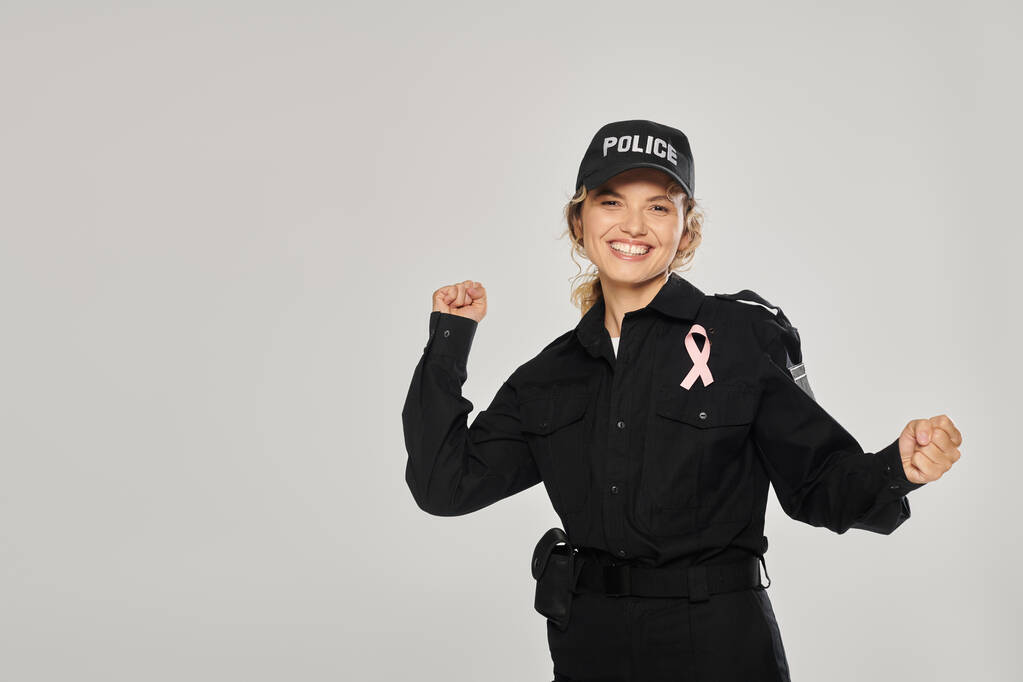 ενθουσιασμένοι αστυνομικός με ροζ κορδέλα του καρκίνου του μαστού δείχνει ναι χειρονομία απομονώνονται σε γκρι - Φωτογραφία, εικόνα
