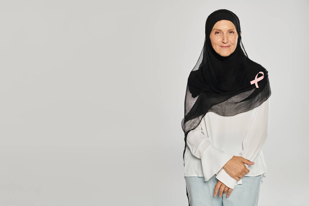 весёлая женщина в хиджабе и розовой ленте осведомлённости о раке молочной железы, стоящая изолированная на сером - Фото, изображение