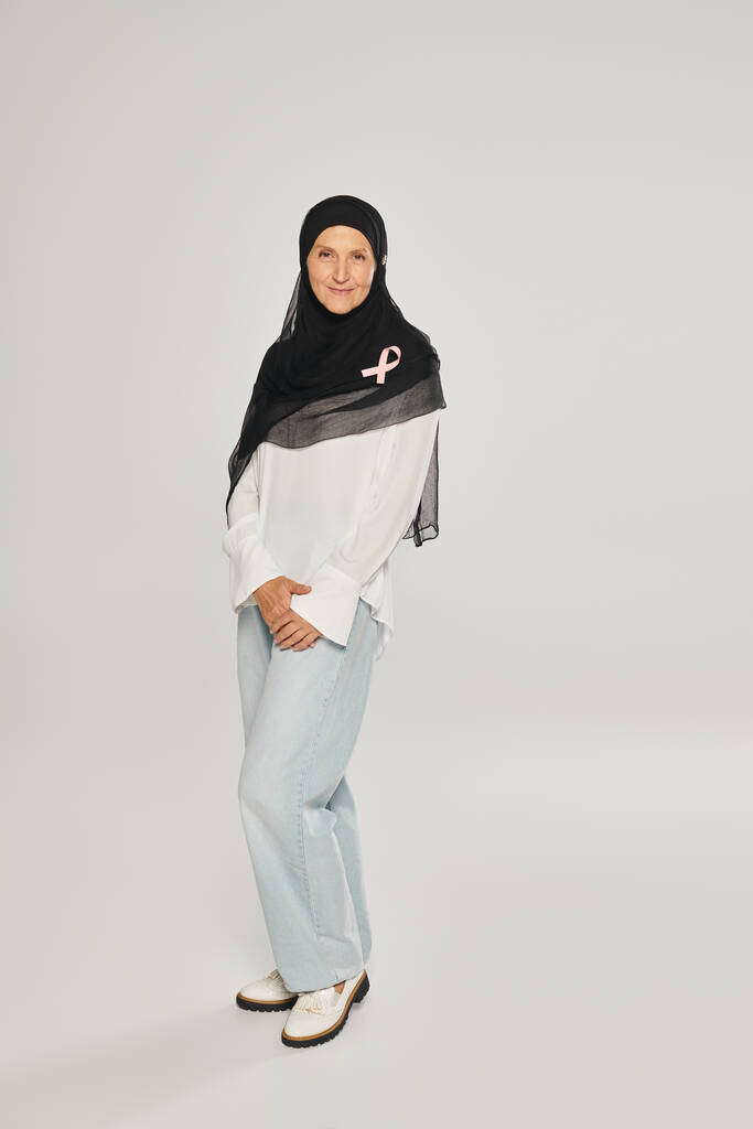 улыбающаяся женщина в хиджабе с розовой лентой осознания рака груди стоя на сером фоне - Фото, изображение