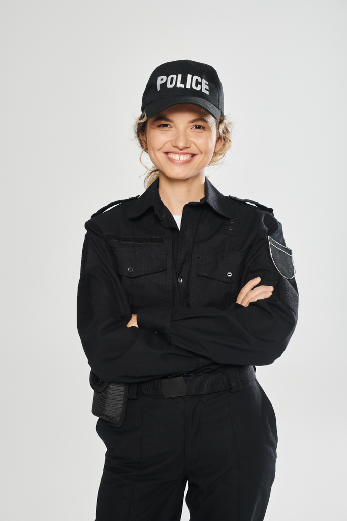 allegra poliziotta in uniforme che incrocia le braccia e posa isolata sul grigio - Foto, immagini