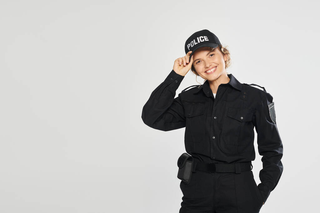 fröhliche Polizistin in Uniform berührt Mütze und blickt in Kamera isoliert auf grau - Foto, Bild