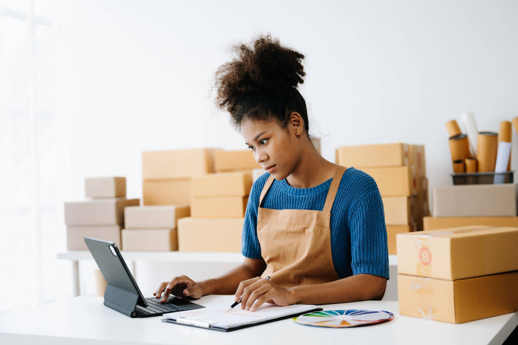 Стартап малого бизнеса МСП, предприниматель владелец африканской женщины с помощью планшетного компьютера, принимая и проверяя онлайн заказ покупки для подготовки упаковки продукта коробка. - Фото, изображение