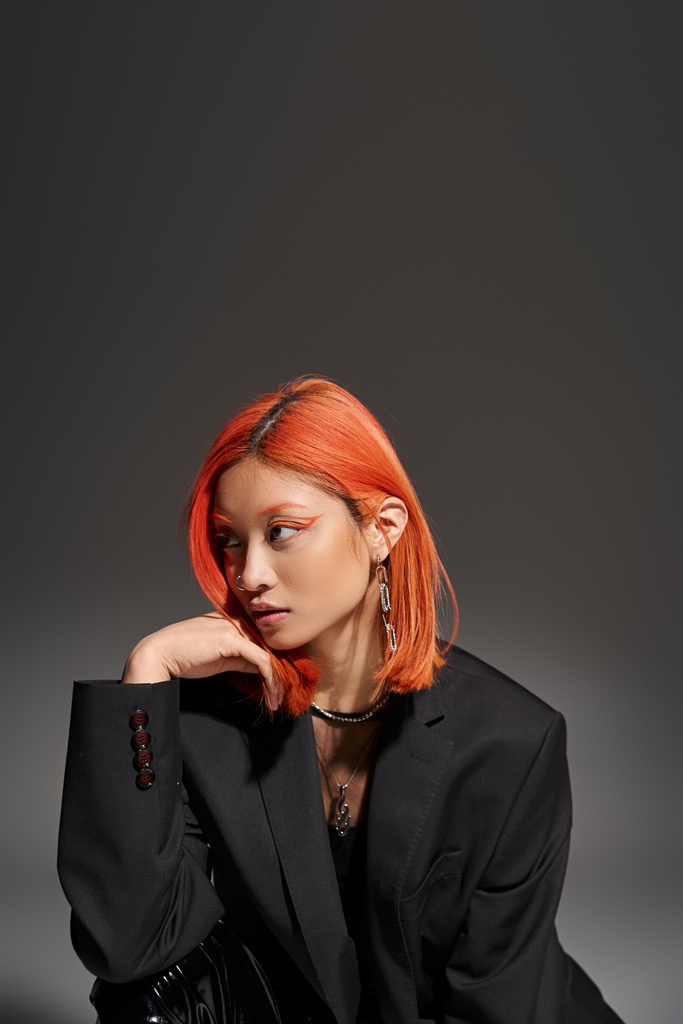 модная азиатка с пирсингом в носу и рыжими волосами позирует в блейзере на сером фоне - Фото, изображение