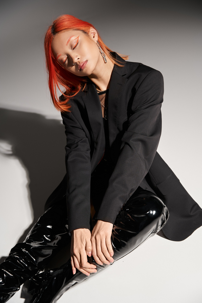 fashionable asian woman in sexy latex boots and blazer sitting with closed eyes on grey backdrop - Photo, Image