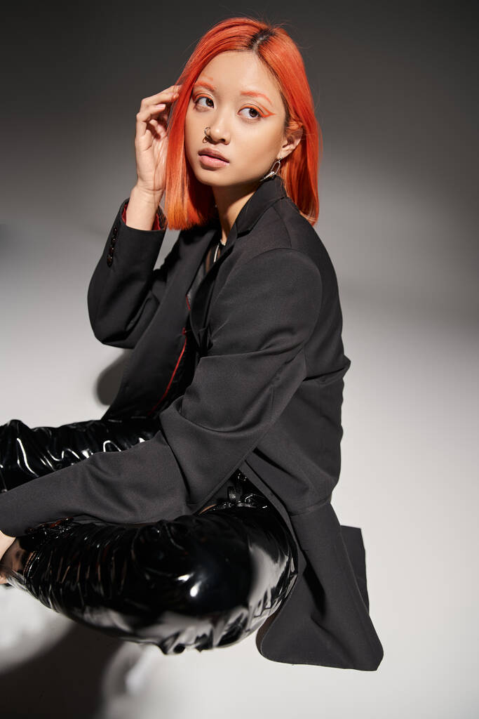 młoda Azjatka z kolczykiem w nosie dopasowując rude włosy i siedząc w odważnych butach lateksowych na szarości - Zdjęcie, obraz
