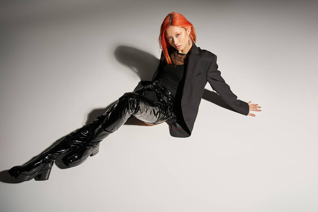 edgy fashion, asian woman with red hair posing in bold outfit on grey background, black latex boots - Photo, Image