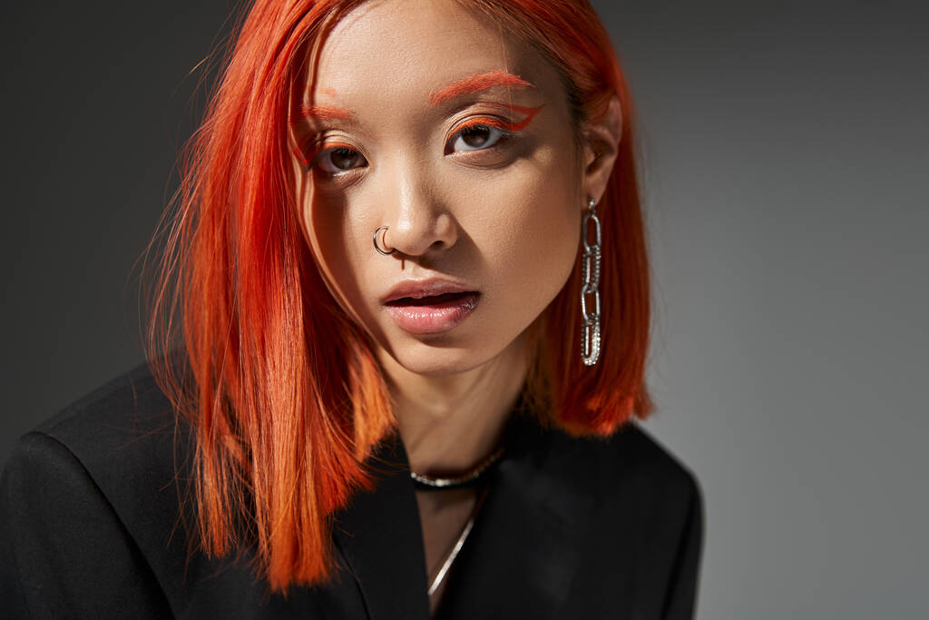 стильная азиатская модель с рыжими волосами и поза пирсинг позируя на сером фоне, портрет - Фото, изображение