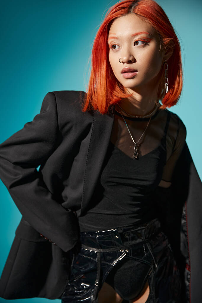 modelo asiático con estilo de pelo rojo y piercing en la nariz posando en chaqueta de gran tamaño sobre fondo azul - Foto, imagen
