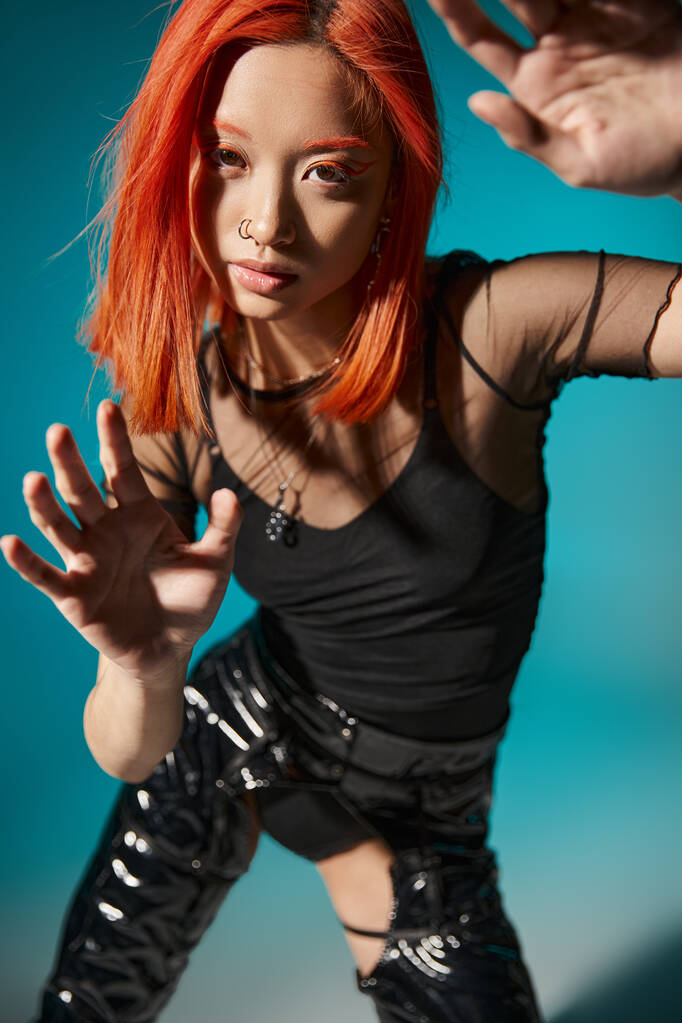 modello asiatico con piercing e capelli rossi tinti gesticolando e guardando la fotocamera su sfondo blu - Foto, immagini