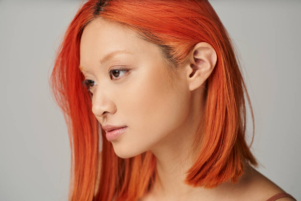 retrato de delicada joven mujer asiática con la piel perfecta y el pelo rojo posando sobre fondo gris - Foto, imagen