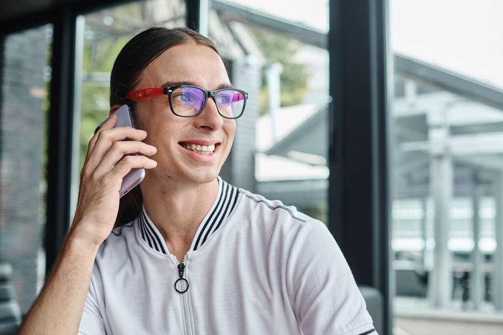 Закрыть веселый молодой человек в очках разговаривает по телефону со стеклянным фоном, коворкинг концепции - Фото, изображение