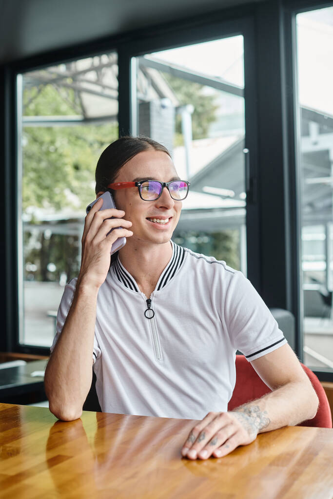 young man with pony tail and glasses talking on phone at table with glass on backdrop, coworking - Photo, Image