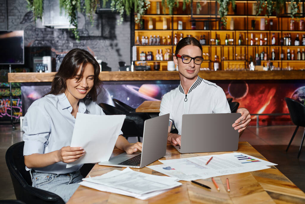 концентрированные улыбающиеся коллеги, работающие над документами с использованием ноутбуков с размытым фоном, коворкинг - Фото, изображение