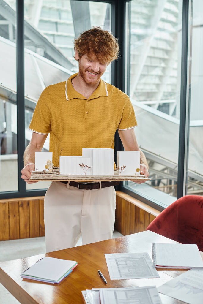 homme aux cheveux roux souriant et tenant modèle d'architecture avec verre sur fond, bureau d'études - Photo, image
