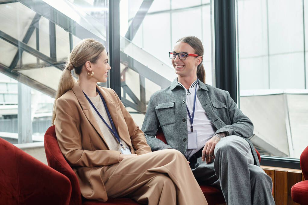 dos elegantes colegas en ropa inteligente sentados en sillas rojas y mirándose, coworking - Foto, Imagen