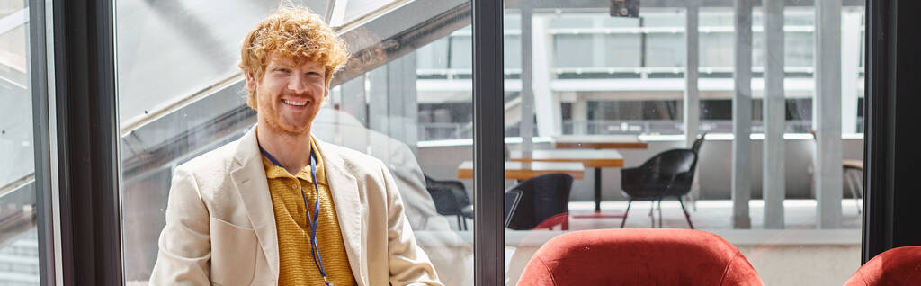 smiling red haired employee in smart outfit looking at camera with glass backdrop, coworking, banner - Photo, Image
