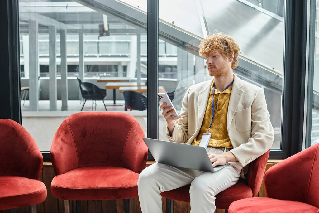 skupiony rudy mężczyzna z laptopem na kolanach siedzi i patrzy na telefon, koncepcja współpracownika - Zdjęcie, obraz