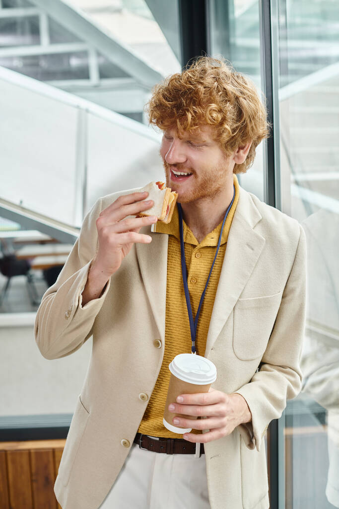 hombre pelirrojo en traje inteligente disfrutando de su café y comer sándwich en la hora del almuerzo, coworking - Foto, Imagen