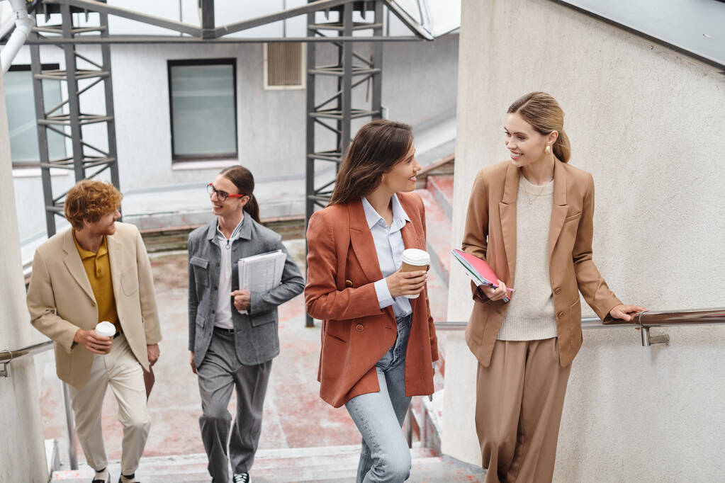 quatro colegas alegres andando lá em cima segurando sua papelada e xícaras de café, conceito de coworking - Foto, Imagem
