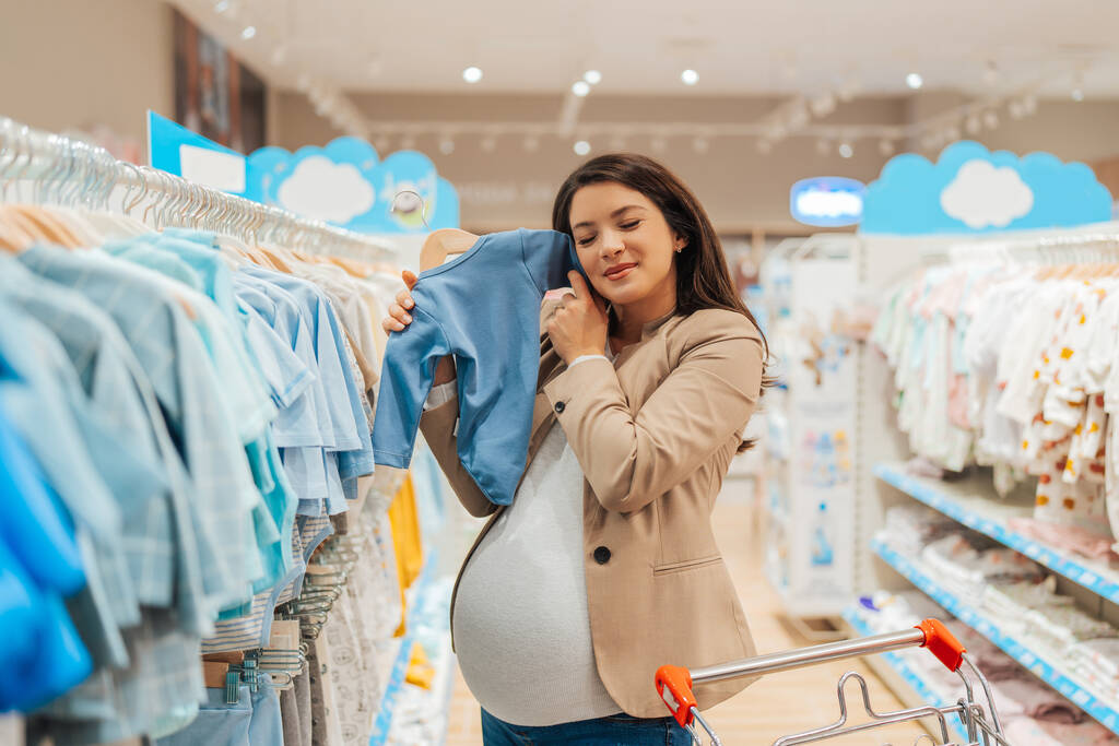 Beautiful young pregnant mother choosing and buying colorful clothes and appliances for her new incoming baby in. Child shop or store concept. - Zdjęcie, obraz