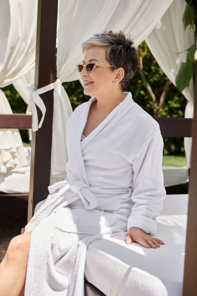 cheerful middle aged woman in sunglasses resting in private beach pavilion during wellness retreat - Photo, Image